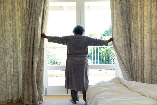 Rear view of african american senior woman opening curtains after waking up in the morning