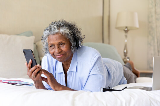 African american senior woman listening music over headphones and using cellphone while lying on bed