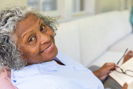 Closeup portrait of smiling african american senior woman with eyeglasses and book relaxing on sofa