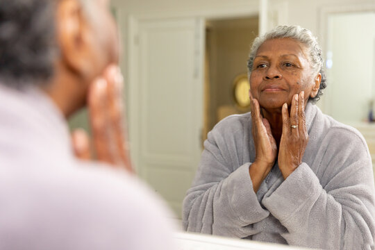 African american senior woman with short gray hair touching her face and looking in mirror at home
