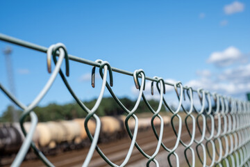 A wire fence separates the freight train. The forest and pale blue sky with white clouds in the distance in the background.