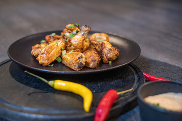 Pieces of grilled chicken with greens on a black plate. Sauce in a black dish and colorful chili peppers