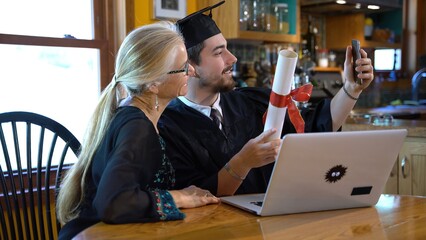 Mother and son sit at dining room table in front of laptop for virtual commencement ceremony and...