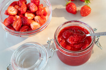 Glass jar with strawberry jam prepared for canning and fresh strawberries in a bowl on the table