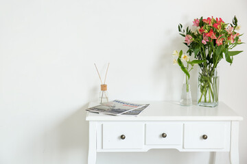 Vases with beautiful alstroemeria flowers, magazines and reed diffuser on table near light wall