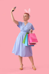 Young woman with bunny ears and shopping bags taking selfie on pink background