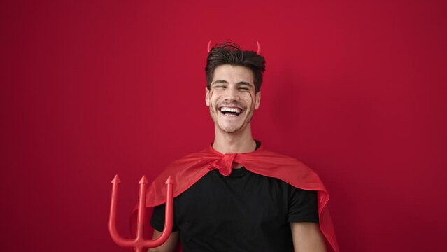 Young hispanic man wearing devil costume holding trident over isolated red background