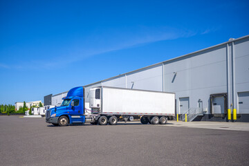 Bright blue day cab big rig semi truck with roof spoiler and refrigerator semi trailer standing in warehouse loading dock waiting for the next load