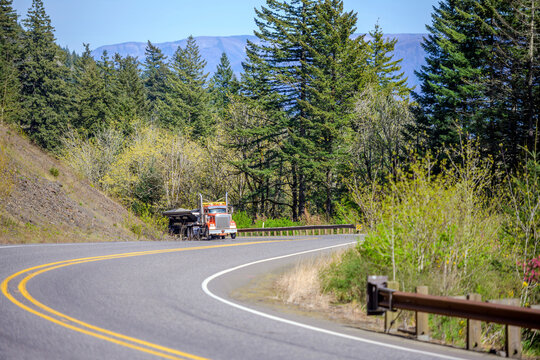 Orange big rig classic semi truck with oversize load sign on the roof transporting loaded tip trailer climbing uphill on the winding mountain highway road in Columbia Gorge