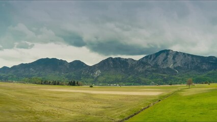 Aerial of lush meadows, towering mountains with rainclouds. The camera moves from the peaceful meadows towards the rugged mountain under a moody sky. Ideal for nature documentaries and travel vlogs.