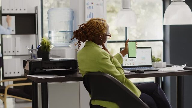 African american woman using greenscreen on phone, working with isolated chroma key display in startup office. Company employee checking blank mockup template on smartphone screen.