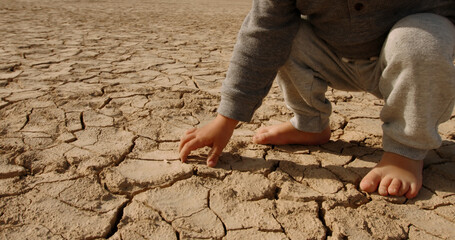 Close up shot of baby boy poking pieces of dry cracked mud. Cracked soil ground of dried lake or...