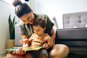 Mom reading a book with baby boy at home. Early age children education, development. Mother and child spending time together. Candid lifestyle. - obrazy, fototapety, plakaty