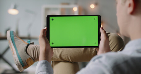 Close up shot of guy lying on couch, having an online video conference, holding tablet computer with mockup chroma key green screen and talking 