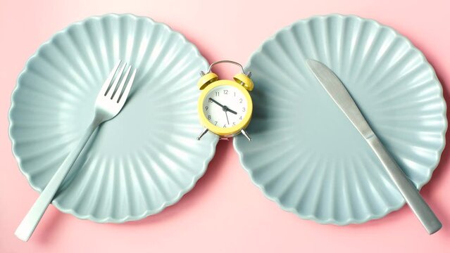Female hand puts an alarm clock on two empty plates on pink background, top view.