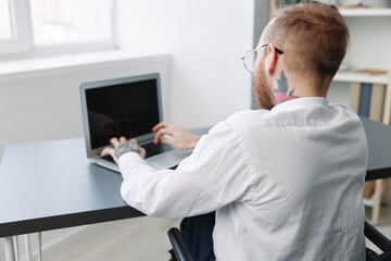Fototapeta premium A man wheelchair businessman with tattoos in the office works at a laptop online, business process, a wheelchair close-up, integration into society, the concept of working a person with disabilities