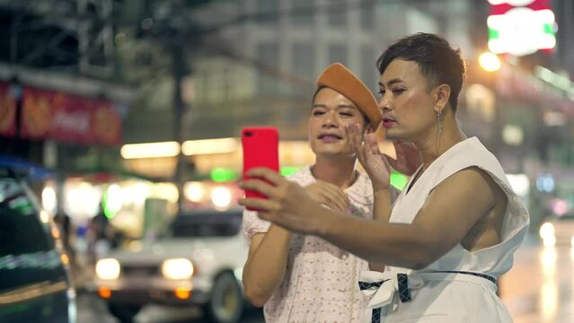 Two Asian guy drag queen in woman clothes and makeup using mobile phone taking selfie together during travel city at night. Sexual equality, lgbtq pride people and transgender cross-dressing concept.