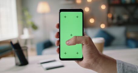 Hand of guys holding a smartphone with green chroma key screen in living room. Template for online store or social media 