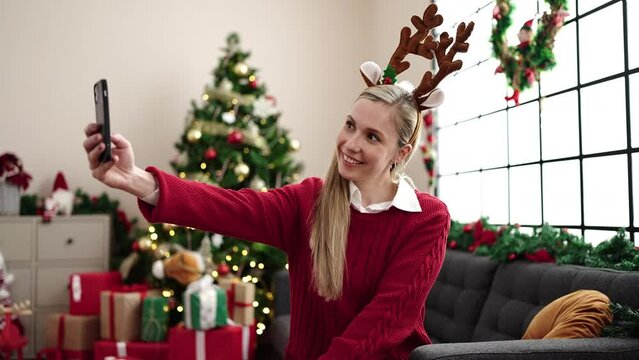 Young blonde woman make selfie by smartphone sitting on sofa by christmas tree at home