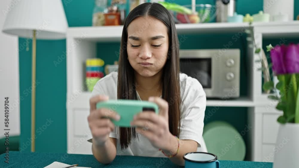 Poster Young beautiful hispanic woman playing video game by smartphone at dinning room