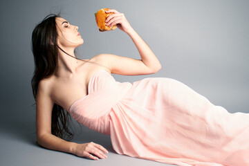 Portrait of woman laying down in ballgown dress pink holding a burger 