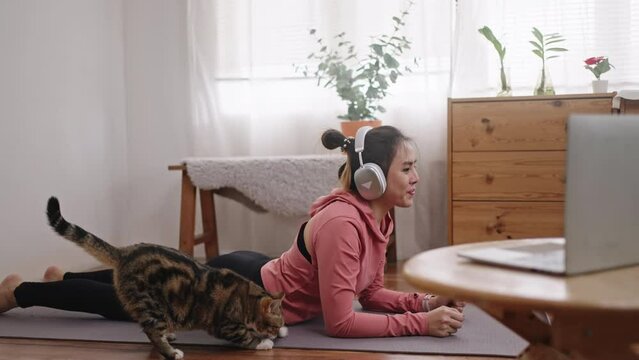 Asian woman in sportswear practicing yoga and watching tutorial lesson on laptop doing exercise in living room with a cat at home.asia girl use headphone in sportswear during yoga.