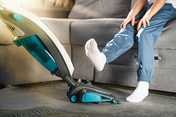 The child raised his legs while sitting on the couch so that his mother could vacuum. House cleaning. Manual vertical vacuum cleaner. Vacuum cleaner is used to clean the carpet in the Living room.