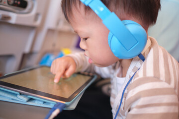 Cute Asian 3 years old toddler boy child wearing headphones using tablet pc watching cartoons, playing game during flight on airplane. Happy Flying with children concept, Soft and selective focus