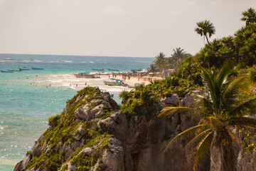 A view of rock cliffs in front of a populated beach blurred out of focus in the background on a sunny summer day with green palm trees and rock and blue ocean waters below.