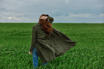 a red-haired woman stands with her back to the camera in a long coat, holding her hand with her hair flying in the wind, standing in a green field in cloudy weather