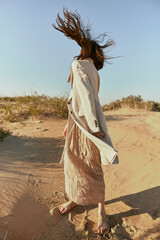 a woman in a light jacket and a skirt blooming sand stands in nature by the sea creatively posing covering her face with hair on a sunny day against a clear sky