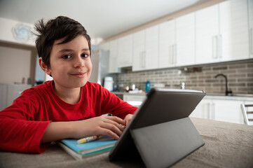 Elementary school student boy on distance learning, smiling looking at camera, doing homework, studying online, watching webinar on digital tablet, holding a pen and making notes in his textbook