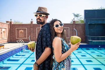 Young Indian travel couple wearing sunglasses and hat drinking coconut water in hot sunny day...