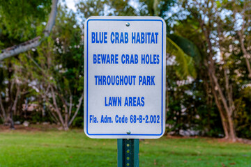 Sign post with Blue Crab Habitat Blue Crab Holes Throughout Park Lawn Areas in Miami, Florida. Close-up of a sign post against the lawn and trees in the blurred background.