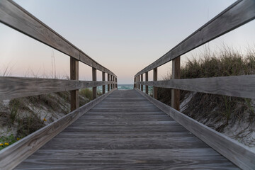 Straight wooden path with railings in between sand dunes at Destin, Florida beach. Walkway with ocean waves below sunset horizon skyline background.