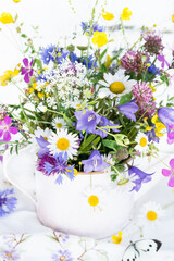 Bouquet of beautiful meadow flowers  in white vase on embroidered tablecloth on white background indoors in natural light, still life with vivid wild colorful flowers, close up view