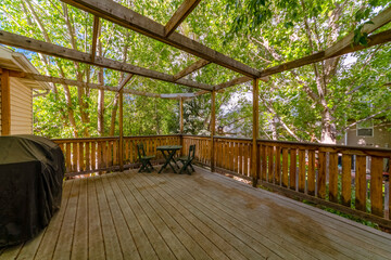 Terrace with wood flooring, railings, and frameworks above. There is a covered grill on the left near the wall with wood lap siding and a table with seats at the corner on the right against the trees.