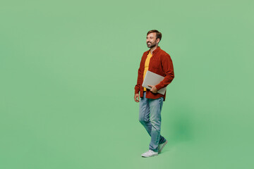 Full body side view smiling elderly IT man 40s years old wears casual clothes red shirt t-shirt hold closed laptop pc computer walk go isolated on plain pastel light green background studio portrait.