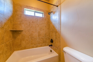 Alcove bathtub with yellowish brown tiles surround in a bathroom with natural lights from the window. Tub shower with wall-mounted showerhead on the left and corner shelf on the right.