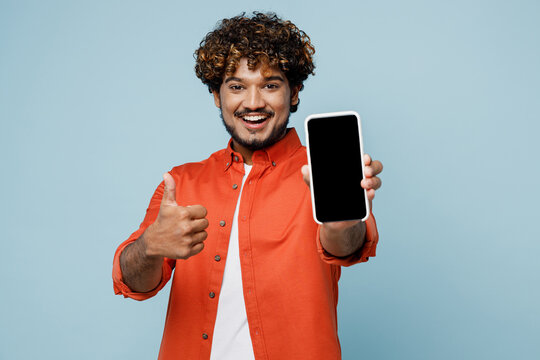 Young fun Indian man wear orange red shirt white t-shirt hold in hand use close up mobile cell phone with blank screen workspace area show thumb up isolated on plain pastel light blue cyan background