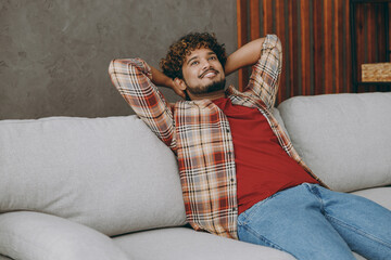 Young smiling calm fun Indian man wearing casual clothes hold hands behind neck sits on grey sofa couch stay at home hotel flat rest relax spend free spare time in living room indoor. Lounge concept.
