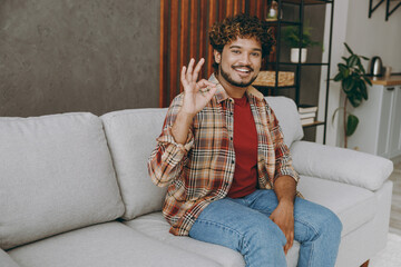 Young smiling happy fun Indian man wears casual clothes showing okay ok gesture sits on grey sofa couch stay at home hotel flat rest relax spend free spare time in living room indoor. Lounge concept.