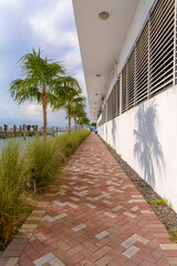 Bricks pavement on walkway beside a building with window blinds panel in Miami, Florida. Vertical shot of a path with grass and palm trees near the water on the left and building with white wall.
