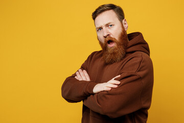 Fototapeta premium Side view shocked sad annoyed displeased young redhead caucasian man wearing brown hoody casual clothes hold hands crossed folded isolated on plain yellow background studio portrait Lifestyle concept