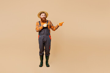 Full body happy young bearded man wears straw hat overalls gumboots work in garden point index finger aside area isolated on plain pastel beige color background studio portrait Plant caring concept