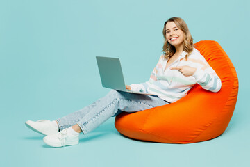 Naklejka premium Full body young IT smiling fun woman wear striped hoody sit in bag chair hold use work point finger on laptop pc computer isolated on plain pastel light blue cyan background studio Lifestyle concept