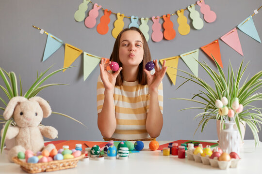 Image of pretty adorable woman with brown hair wearing striped shirt celebrating Easter, holding colored eggs, sending air kissing, congratulating with holiday against decorated festive wall