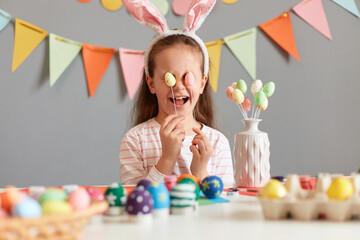 Portrait of happy funny little girl wearing rabbit ears sitting at table having fun while preparing...