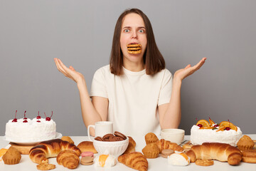 Portrait of shocked confused uncertain woman with brown hair wearing white T-shirt sitting at table isolated over gray background, shrugging shoulders, overeating sweets, has full mouth of cookies.