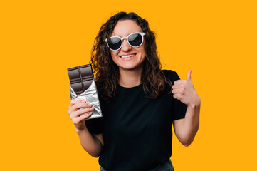 Handsome woman wearing glasses is holding chocolate bar and a thumb up.